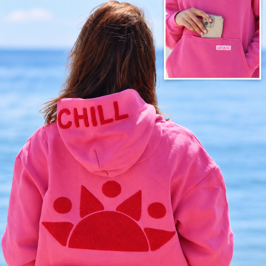 A woman standing on a beach with her back to the camera, wearing a bright pink hoodie with 'CHILL' printed in red across the hood and a large red sunburst graphic on the back. An inset in the top right shows a close-up of the hoodie pocket holding a phone.please size this so it fits appropriately in the block and make sure to keep the inserted image locatedin the top right hand corner